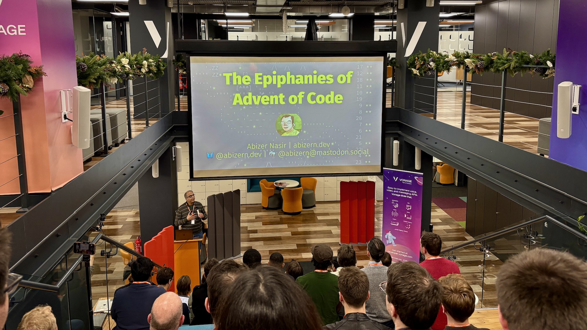 Photo of people attending talk. Slides show “The Epiphanies of Advent of Code”, “Abizer Nasir”, “abizern.dev”.