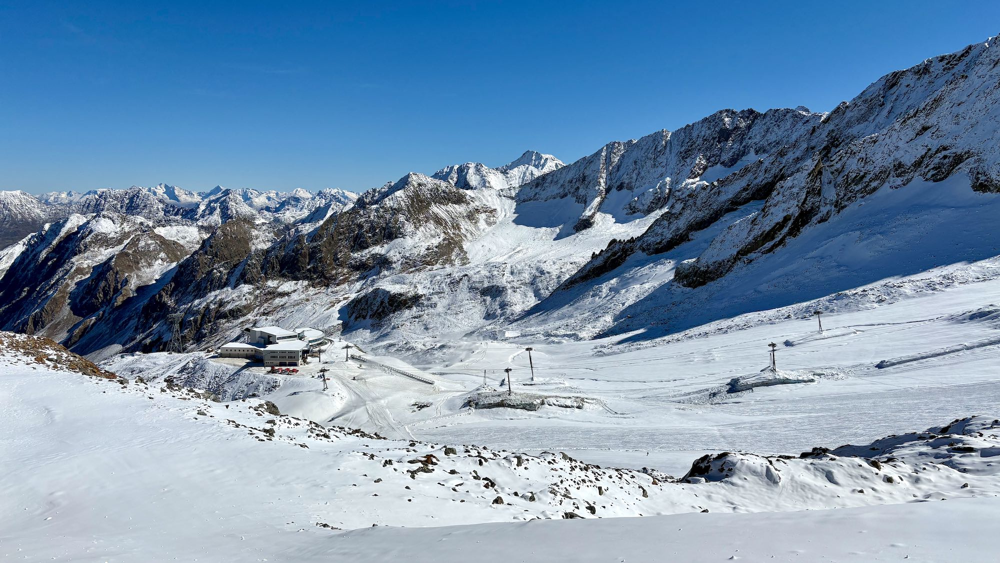 Photo of snow on mountains with ski runs and lifts. Rocky ground not covered with snow in places. Blue sky above.