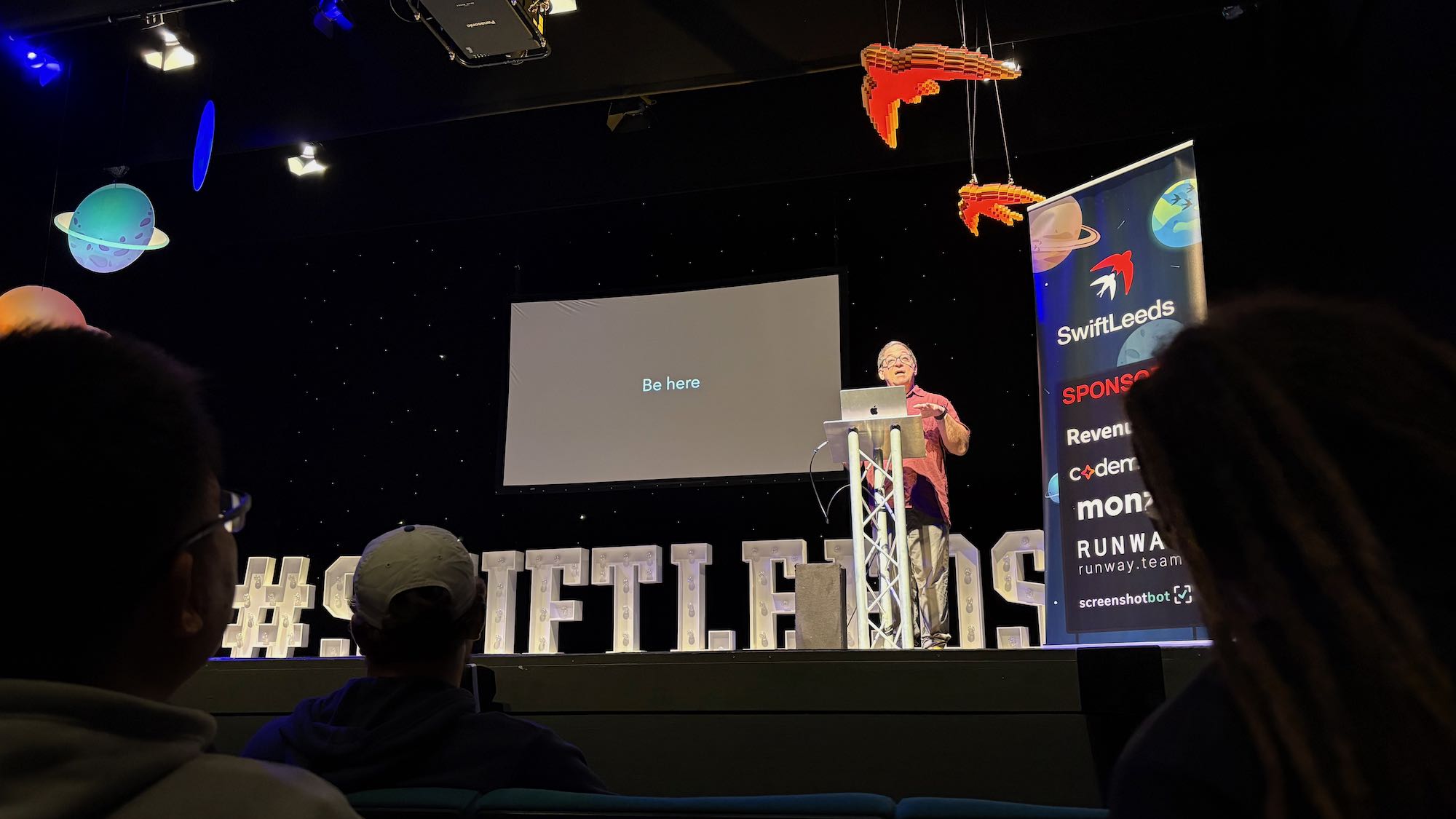 Photo of Daniel on stage at the conference. Slide says “Be here”.
