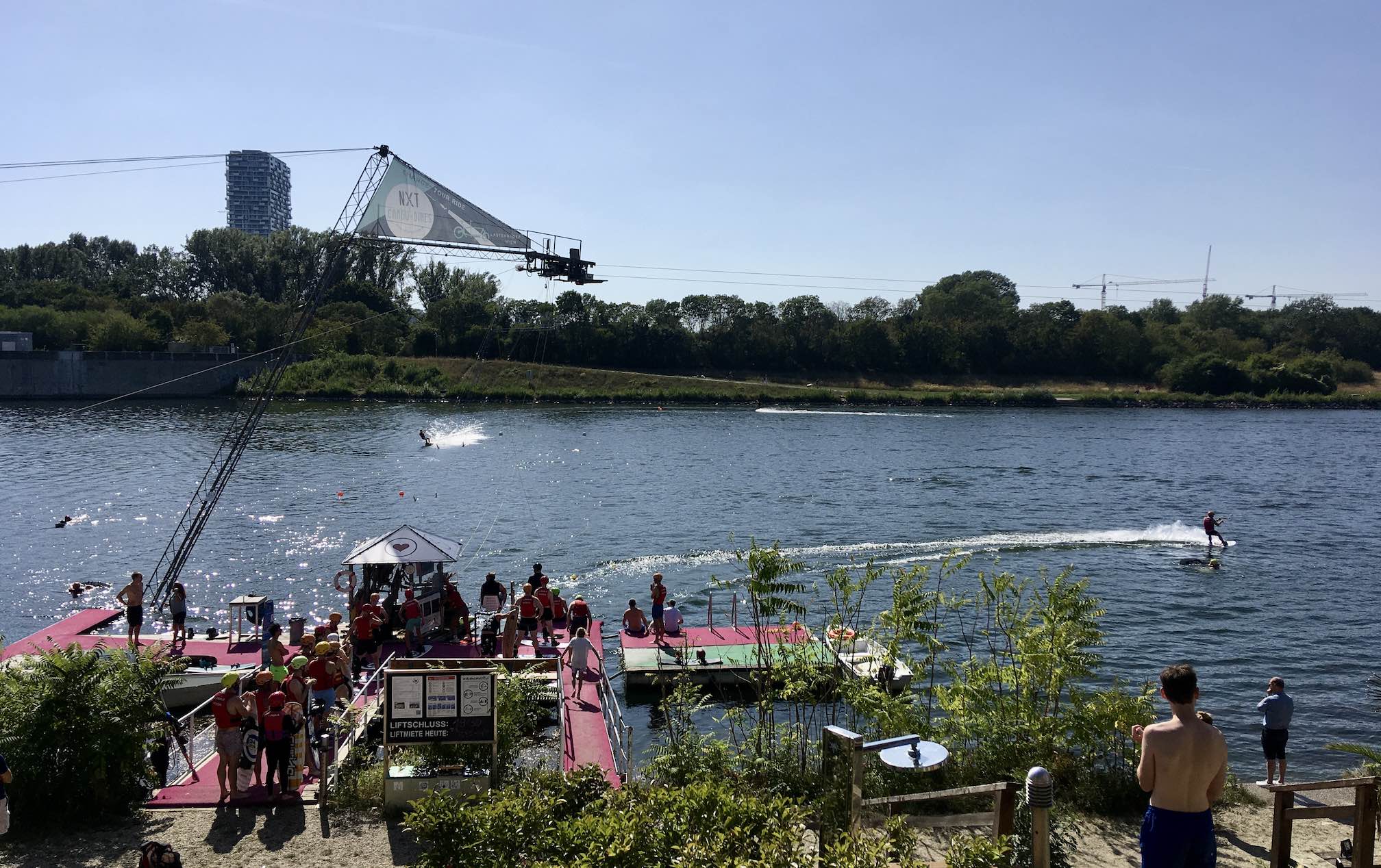 Sunny photo of cable wakeboard park by river with a queue of people on the dock